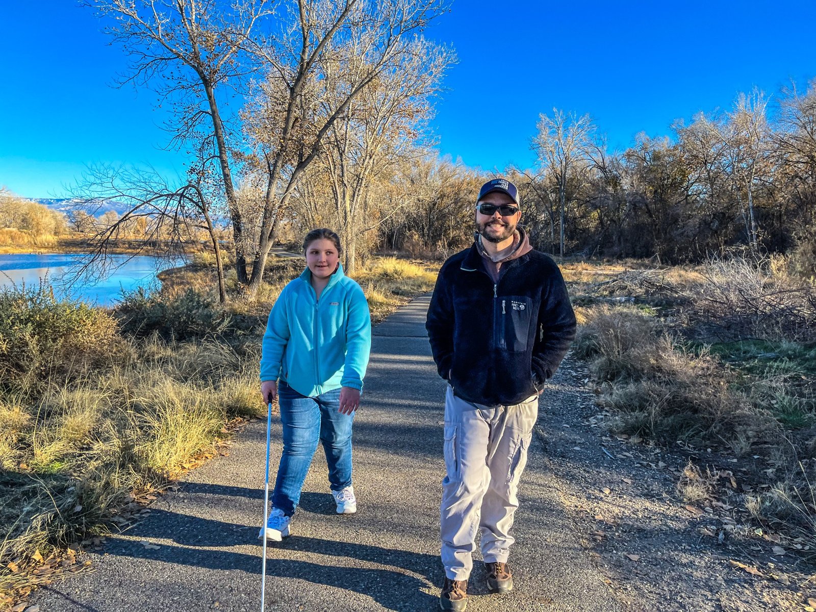 Rob and Isabelle Harris walk beside the water on a paved trail; Isabelle uses her white cane.